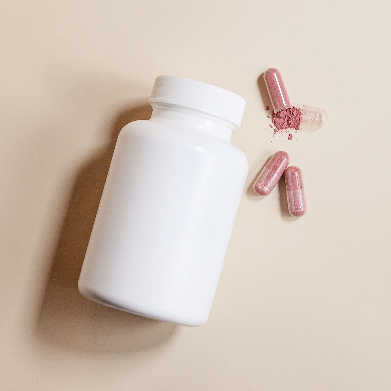High-angle view of a white supplement bottle with pink capsules on a beige surface.